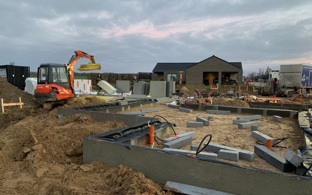 Byggeplads i skumringen med gravemaskine, sokkelblokke og nybyggede huse i baggrunden